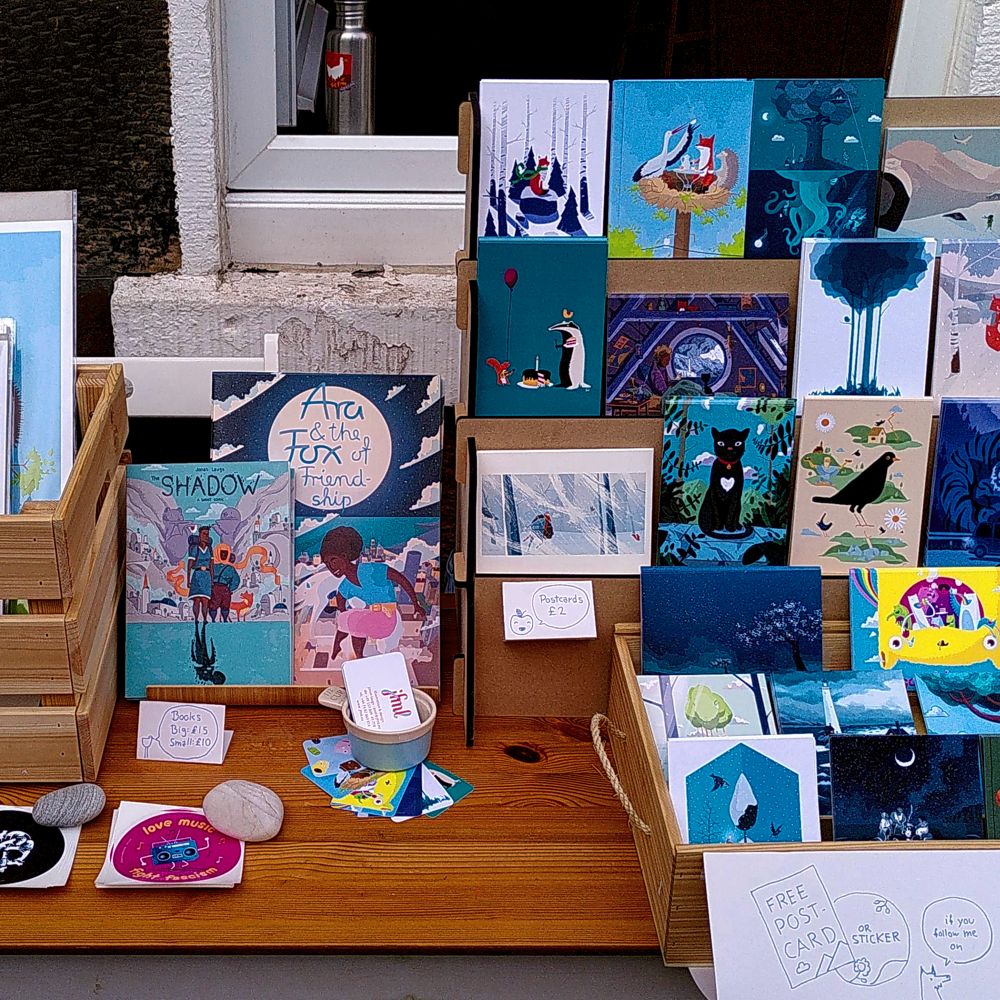 Photo of a table with lots of my postcards, books, stickers, business cards and prints