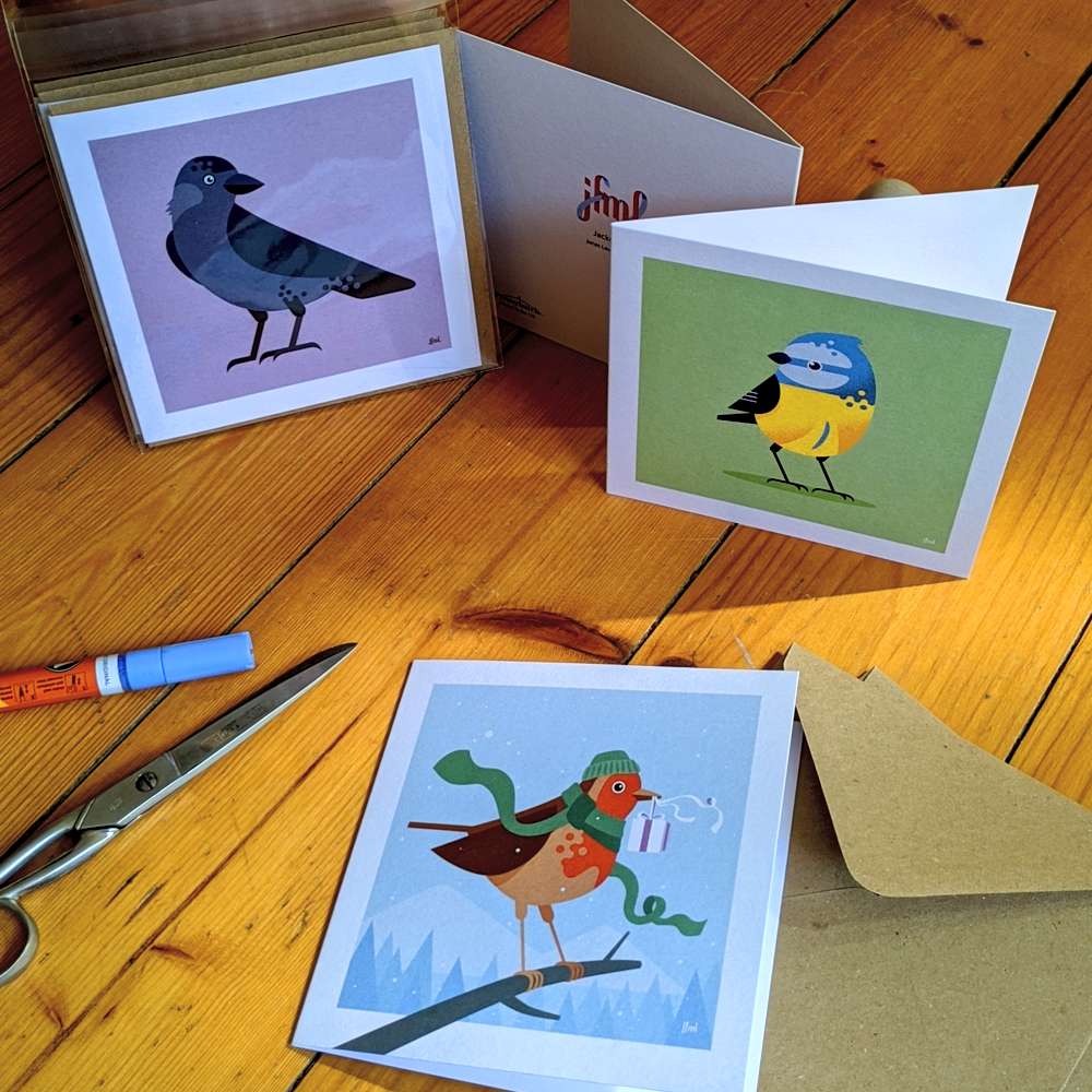 Photo of three square greeting cards with birds on them, on a wooden floor.
