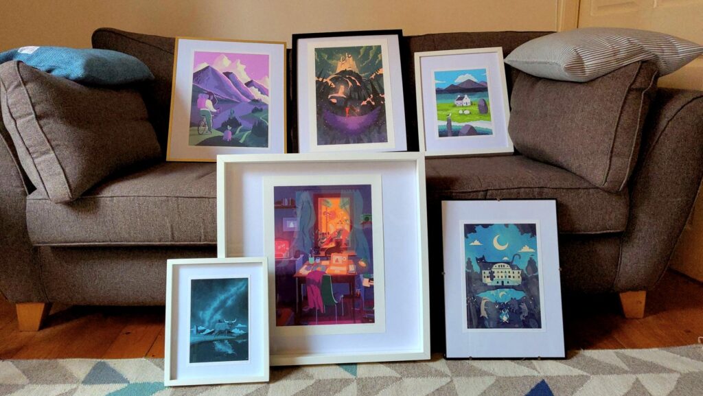 A bunch of framed prints arranged on a grey sofa, the prints feature mostly landscapes in blue and pink.