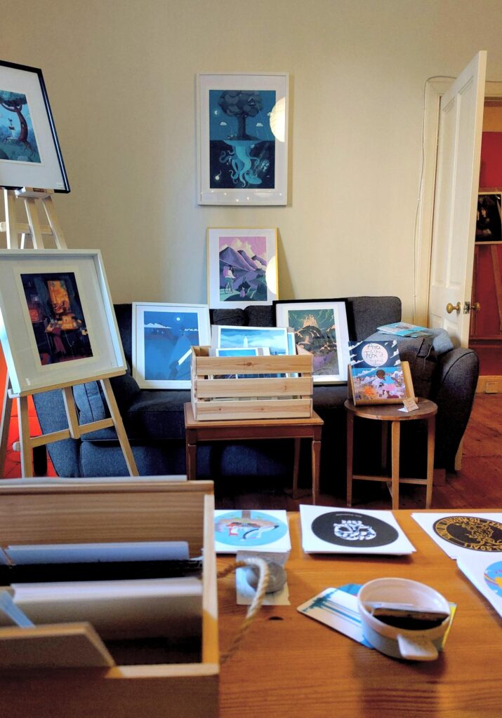 My POV at the Colony of Artists: a table in the foreground with postcards and stickers on them looking out into a room with prints in frames on an easel and on a sofa, there's two small tables with prints and comic / children's books as well.