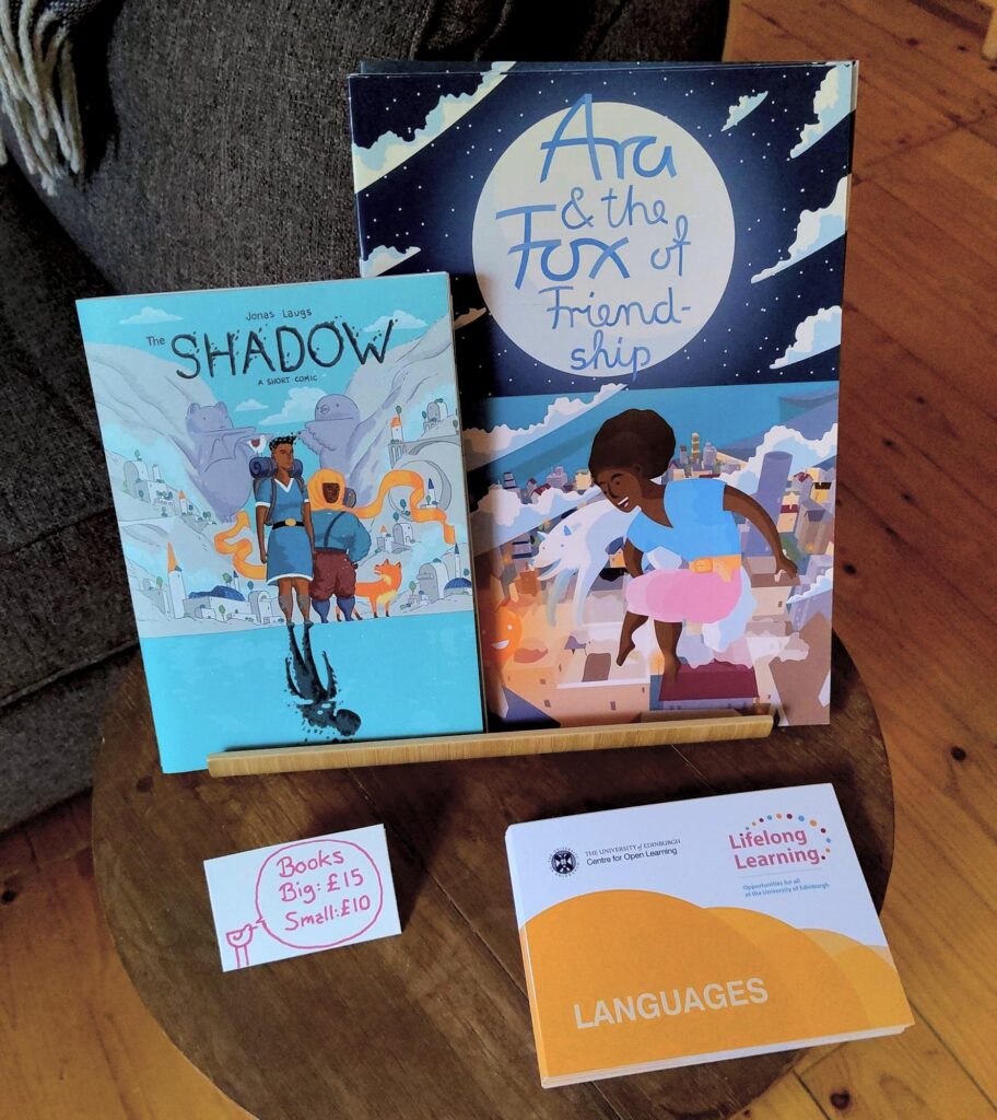 Close-up of the table with the comic (The Shadow, two people standing by a lake in front of a ruined city on a mountainside) and children's book (Fox of Friendship, a girl and a fox flying above a nightly city) from the last picture.